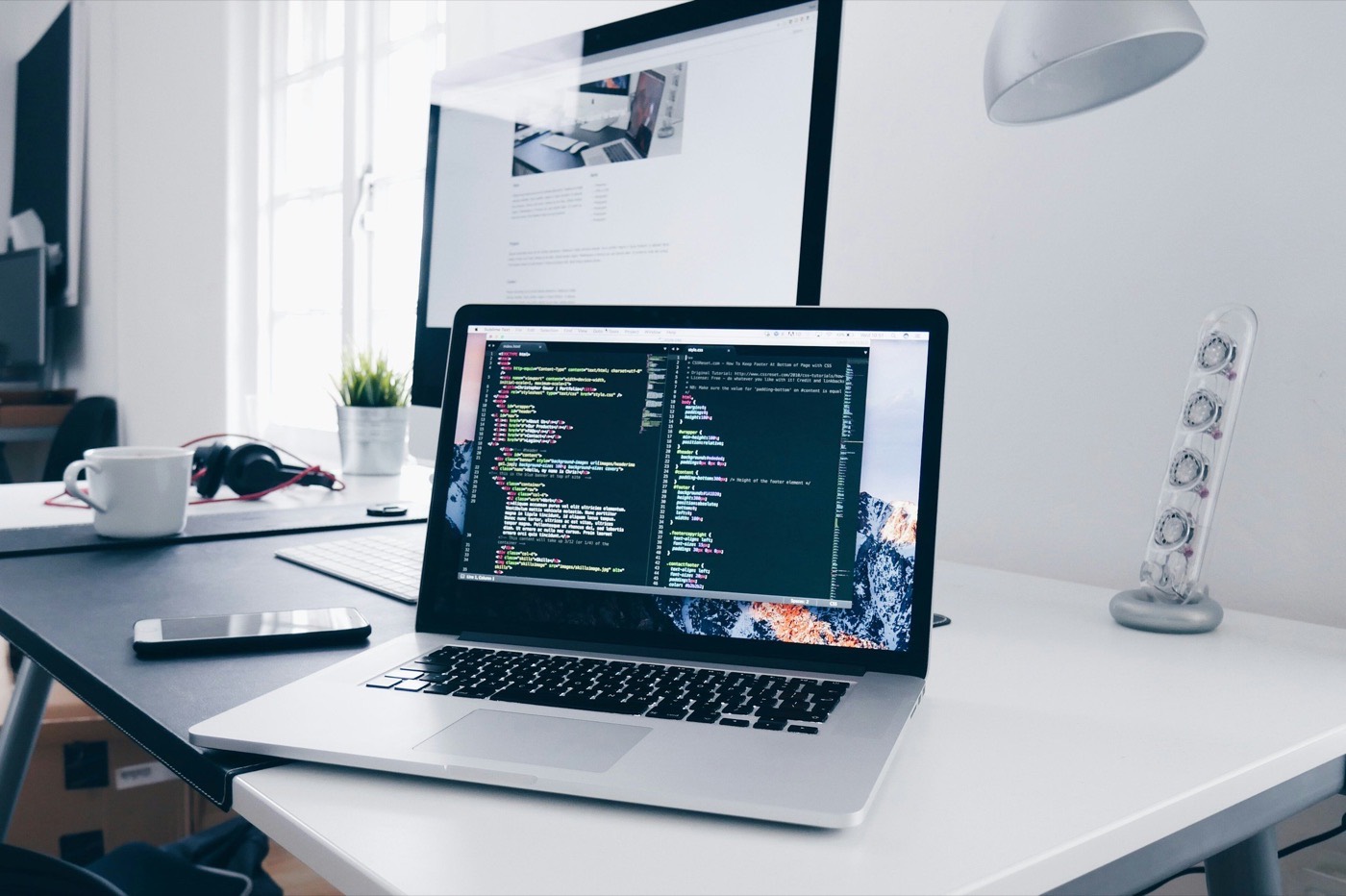 Laptop showing code editor beside a monitor on a bright minimalist desk
